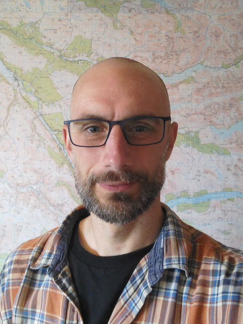 DR with glasses, stubble and balding.  Wearing a black tshirt and checked shirt.  Against a background picture of a map